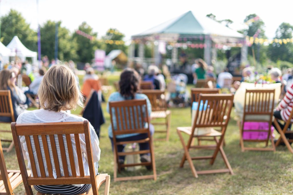 People sitting in foldable chairs watching a band play on a bandstand on a sunny day.