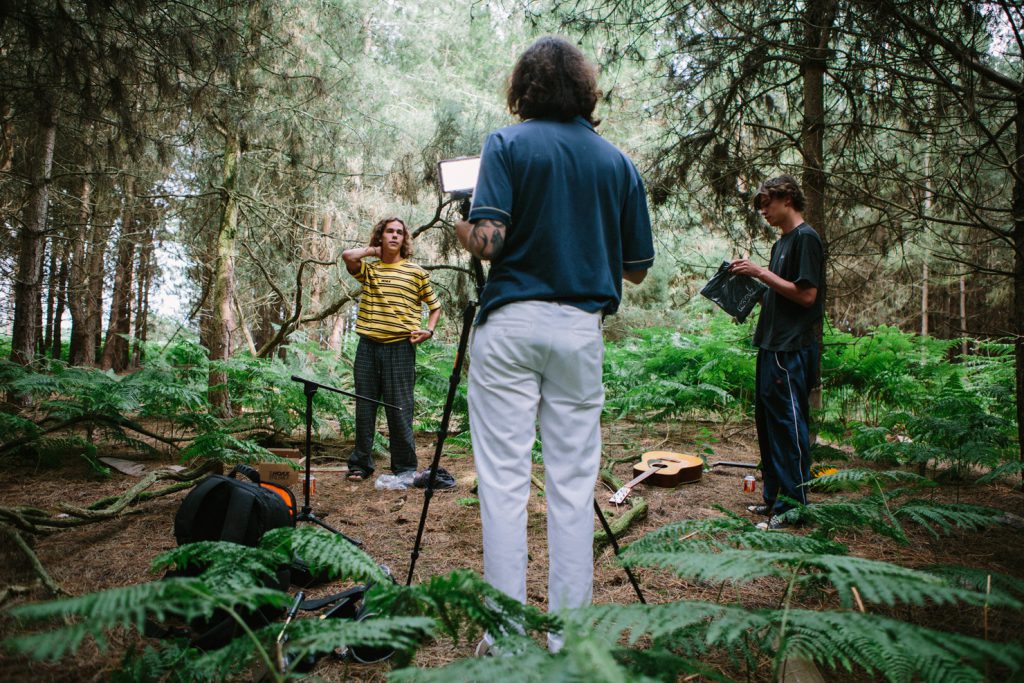 Ricardo Markin standing with a film camera on a tripod with his back facing towards the camera with two young men in front of him in the woods. There is also music equipment on the floor and a guitar.