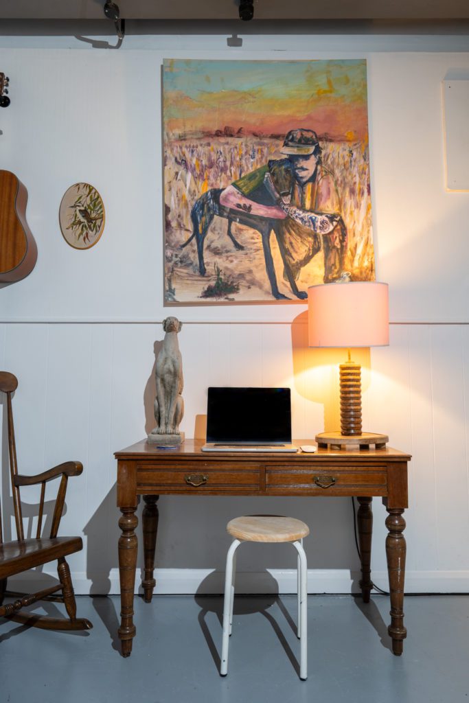 Picture of an old style desk with a lamp and laptop on top as a painting sits above it on the wall.